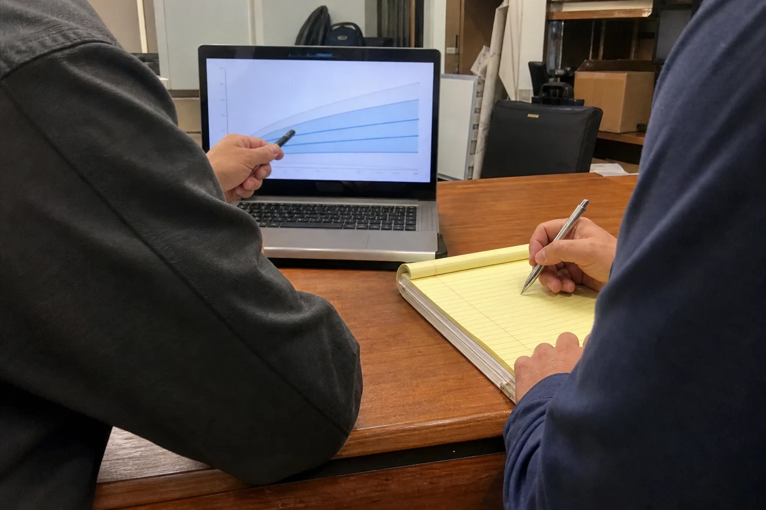 Over-the-shoulder view of two people at a wooden office desk, one pointing a pen at a growth chart on a laptop while the other writes on a yellow legal pad