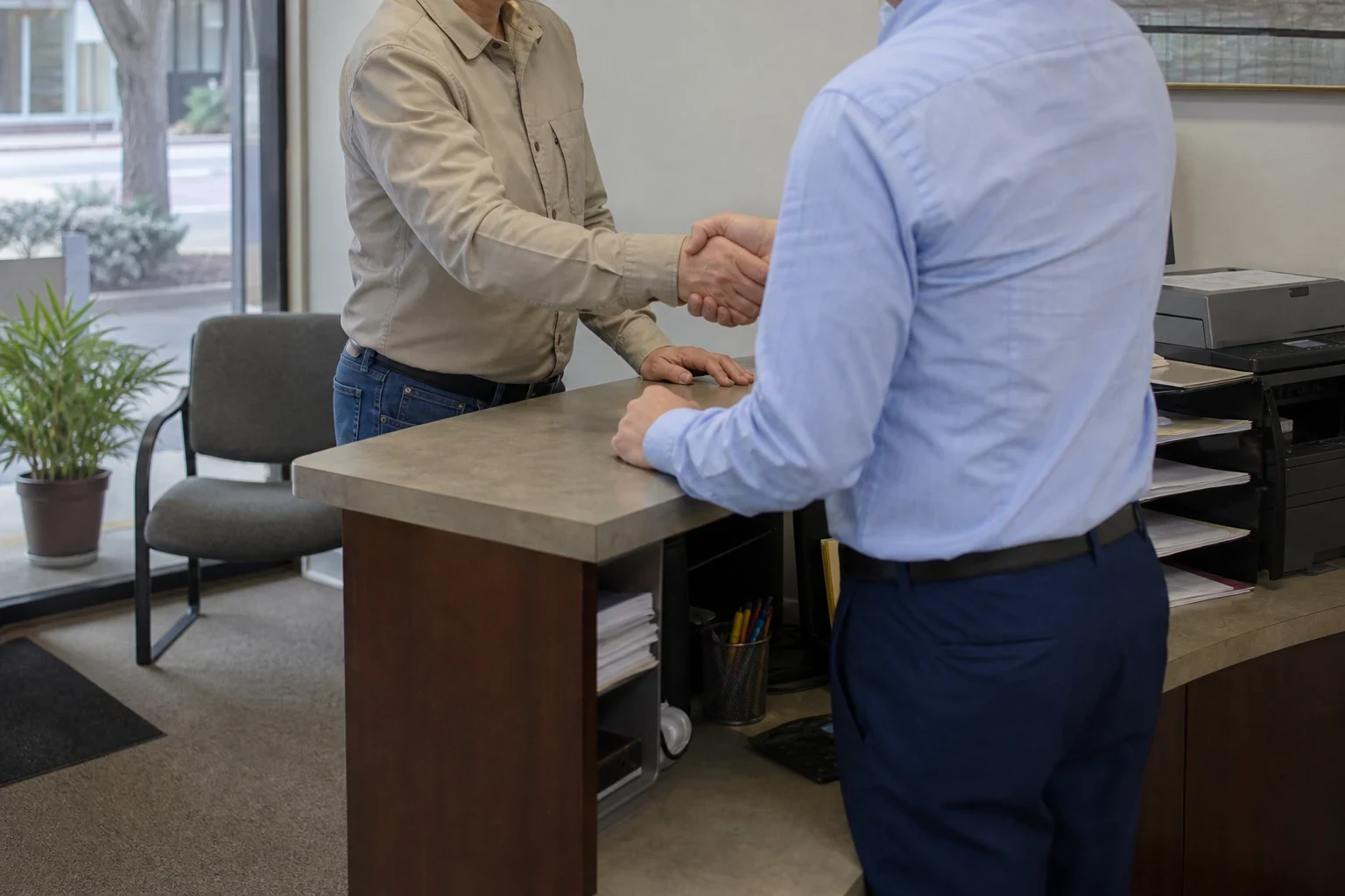 Agent in a light blue shirt shakes hands with a client in a khaki shirt across a reception counter, with a waiting chair and potted plant by the front window