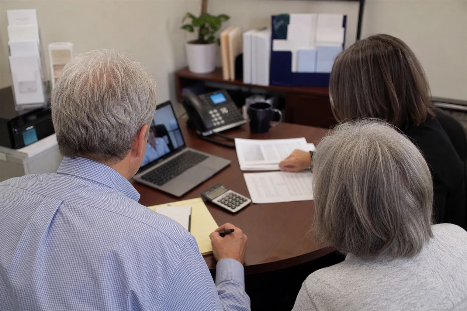 Gray-haired couple seen from behind at a round office desk with an agent reviewing papers, the man taking notes on a legal pad beside a calculator and laptop